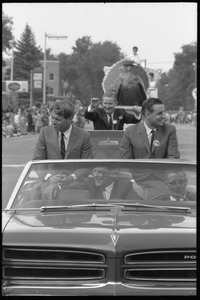 Robert F. Kennedy (left) and Walter Mondale riding in an open car at the Turkey Day parade while stumping for Democratic candidates in the northern Midwest (giant turkey float visible in the background)