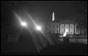 Nighttime view of a row of tents, one with a sign reading 'Houses not tents,' the White House and Washington Monument in the background