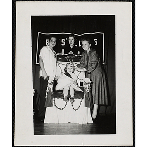 The Little Sister Contest winner, sitting on her throne, receives a crown from Barbara Sherman Burger while her brother and an unidentified woman pose behind her