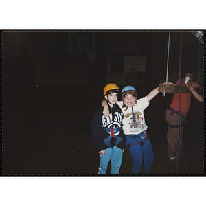 Two girls wear safety harnesses and helmets in a gymnasium