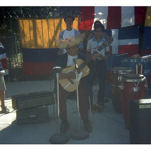Male, Hispanic American musician wearing a sombrero plays a guitar at a Latino street festival; on the right, a boy plays maracas
