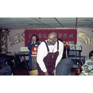 Mel King takes a seat at a restaurant table near a podium where a woman addresses the group gathered for a celebration of the Chinese New Year