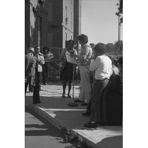 Two women stand at a microphone and look at a large sheet of paper, with onlookers listening at a Latino street festival
