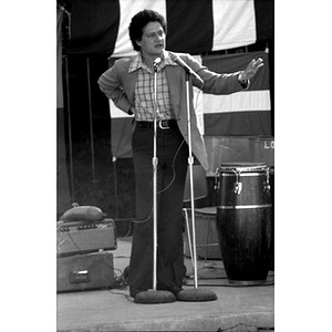 Hispanic American man speaks into two microphones at a Latino festival; he is standing and gesturing to the right with his left hand outstretched