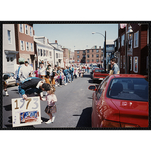 A man holds a parade sign reading "1973 PICASSO" and bends down to talk to a girl while a pipe band walks by during the Boys and Girls Clubs of Boston 100th Anniversary Celebration parade