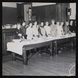 Women stand behind tables of baked goods during a Mothers' Club Cake Walk event