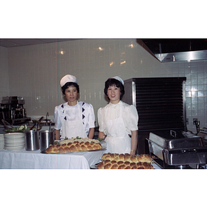 Two women in white aprons and hats stand behind a food service station laden with bread loaves and salad