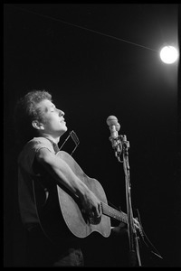 Bob Dylan, with guitar and harmonica, performing on stage, Newport Folk Festival