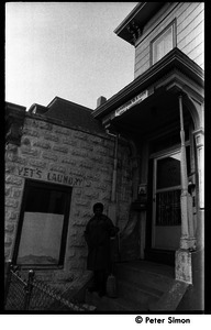 African American woman with broom standing on the front steps of 199 Harvard St., Cambridge, Mass.; Vet's Laundry sign in background and banner over doorway reads 'Cambridge is a city, not a highway'