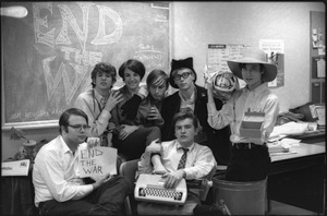 At the Boston University News Office: staff of BU News, Don McClean holding sign, Peter Simon top left, unidentified woman, unidentified man, Clif Garboden, Ed Siegel, and Joe Pilati in front with typewriter, December 1967