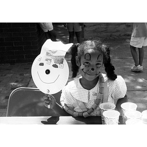 A little girl with her face painted displays the art project she has made.