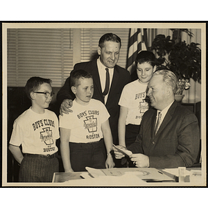 Mayor of Boston John F. Collins sits at his desk and smiles at a group of three boys wearing Boys' Clubs of Boston t-shirts while William J. Lynch stands behind them