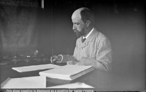 Henry Adams seated at desk in study, writing, in light coat