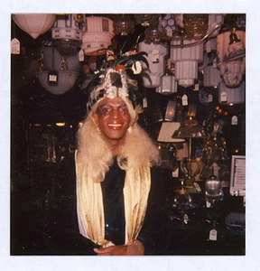 A Photograph of Marsha P. Johnson with Blonde Hair, a Black Bejeweled Headpiece and Gold Scarf at Uplift Lighting