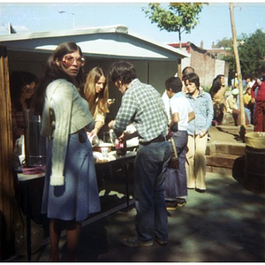 Three people stand in line to buy food at a vending stand at a Latino street festival, with at least two people selling the food, and several people standing nearby and in the (right) background
