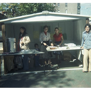 Three woman and a small boy sell food at a vending stand at a Latino street festival; a woman stands in front of the table, on the right