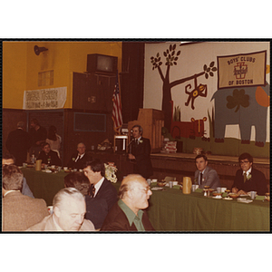 A man with a carnation speaking from the podium at the head table during a Boys' Club St. Patrick's Day event