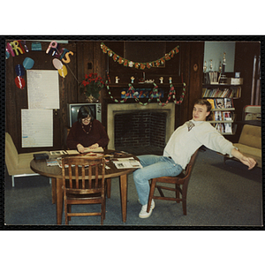 A woman and a teenage boy sit a table with "Merry Christmas" banner hanging over a mantle