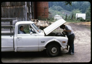 Tony Mathews looking under the hood of pickup truck by the barn, his mother in the passender seat, Montague Farm Commune
