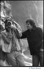 Rameshwar Das taking a photograph, standing with Peter Simon by a granite outcropping on the beach