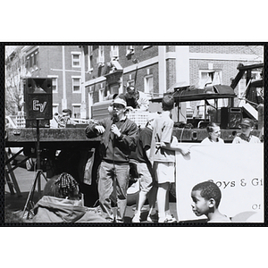 Jerry Steimel, Charlestown Clubhouse director, speaking into a microphone while several boys hold a Boys and Girls Club banner in front of a mobile stage truck at the Boys and Girls Clubs of Boston 100th Anniversary Celebration event