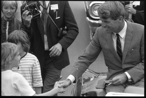 Robert F. Kennedy seated in an open car in a parade, shaking hands with a youngster while stumping for Democratic candidates in the northern Midwest