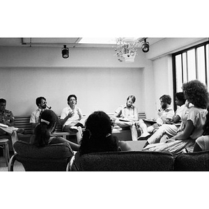 Group of people sitting around the edges of a room listening to an unidentified man and taking notes.