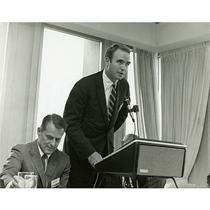 Henry R. Guild, Jr. speaks at the podium while Richard Harte, Jr. sits beside him