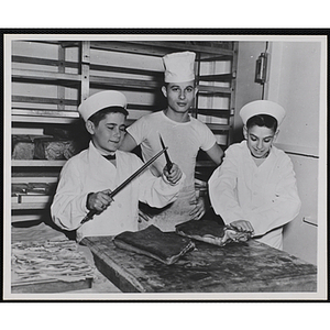 Two members of the Tom Pappas Chefs' Club sharpen a knife and work with cuts of meat under the supervision of a sailor aboard the USS Salem