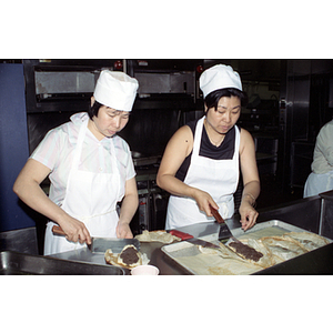 Two women wearing white aprons and hats work in an industrial kitchen