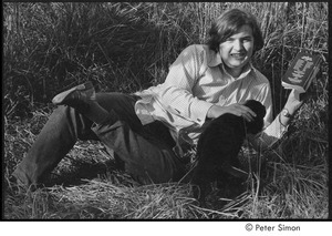 Joe Pilati in tall grass, holding a book and petting a puppy, Packer Corners commune