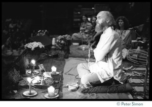 Ram Dass seated onstage at the Winterland Ballroom during his 'marathon,' looking out toward the audience, arms crossed