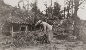 Red Cross worker [Elsa Bowman] picks up some debris in front of a hillside dugout, Pinon
