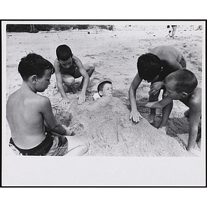 Four boys cover another boy with sand up to his neck on a beach