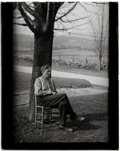 Robert Frost, seated beneath a tree, stone walls and fields in the background