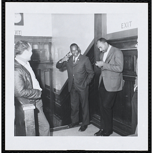 A man puts a starting pistol to his head as another man holds a stop watch and a third man looks on in a building