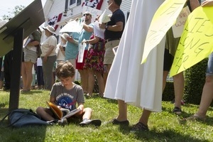 Young boy at a pro-immigration rally in front of the Chatham town offices building, seated on the grass, reading a book : taken at the 'Families Belong Together' protest against the Trump administration's immigration policies