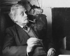 Hugh MacDiarmid, sitting in chair smoking a pipe, just extinguished match