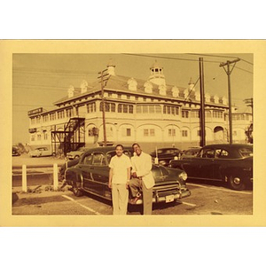 Two men lean against a Chevrolet in a parking lot