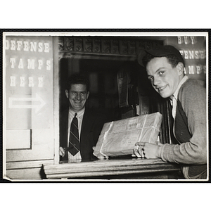 A teenage boy posing with a package at the post office while a man smiles behind the counter