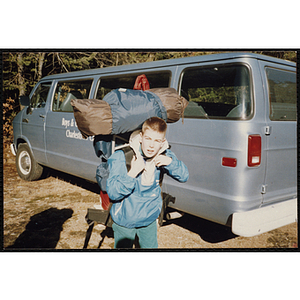 A boy carries a large backpack next to a van outdoors
