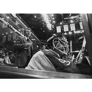 A hockey player sits on the bench as a coach stands behind him watching the game