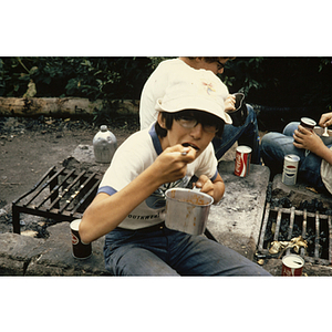 Seated boy eating from metal pot along outdoor fire pits
