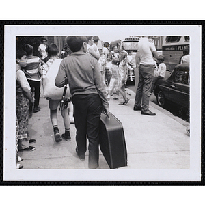 A boy walks with a suitcase and fishing rod as buses wait on the street