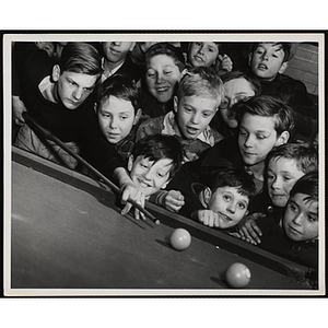 A Large group of boys crowd around a pool player as he prepares to take his shot