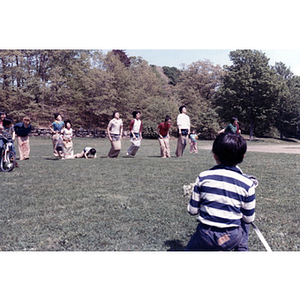 Association members compete in a sack race in a grassy field, while a young boy watches from the sideline