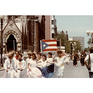 A group marches on a street wearing white and carrying a Puerto Rican flag