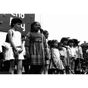 Girl at the microphone on an outdoor stage along with other children.