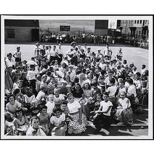 A large group of girls and boys pose for a group shot with a man playing a guitar on a playground during a day camp square dance