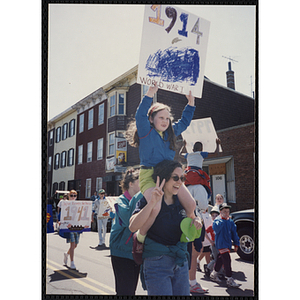 A girl holds up a sign reading "1914 World War I" while riding on a woman's shoulders at the Boys and Girls Clubs of Boston 100th Anniversary Celebration Parade